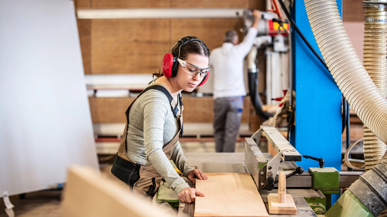 Eine Schreinerin mit Gehörschutz und Schutzbrille arbeitet in einer Werkstatt mit Holz.